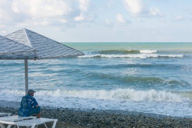 Yalnız adam kumsaldaki beyaz iskemlede oturur ve büyük dalgalara bakar..