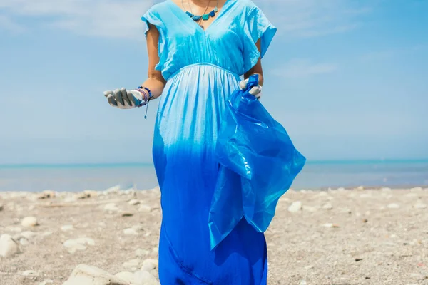 Young woman activist on the beach wears gloves and a garbage bag, wears a blue dress. Earth day concept.