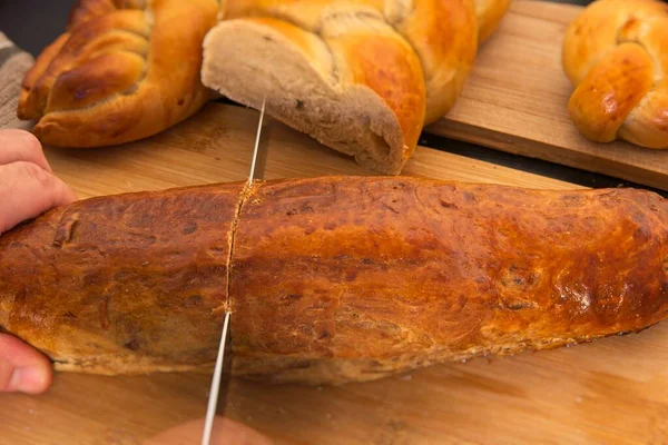 Osoba z nożem krojącym bochenek chleba. Chleb w kształcie warkocza. Słodka Chała. śniadanie, widok z boku. Pomidory wiśniowe i oliwa z oliwek — Zdjęcie stockowe