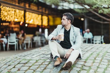 Parkta oturan bir iş adamı elinde kahve ve kafeteryanın sarı ışıklarının olduğu bir arka plan.