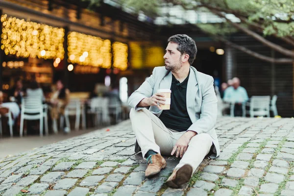 Parkta oturan bir iş adamı elinde kahve ve kafeteryanın sarı ışıklarının olduğu bir arka plan.