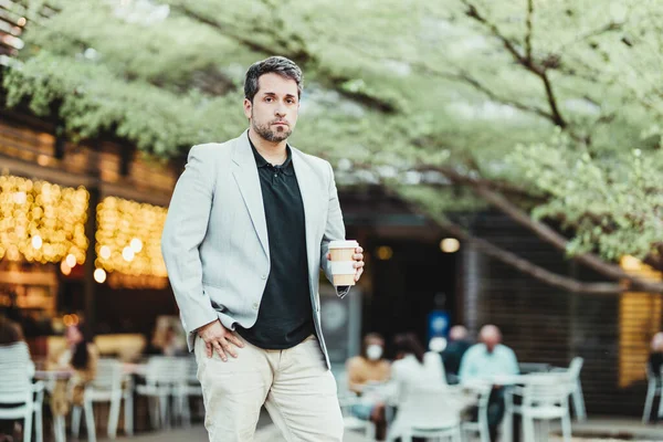 Parktaki bir iş adamı elinde kahveyle kameraya ve kafeteryanın sarı ışıklarının arka planına bakıyor.