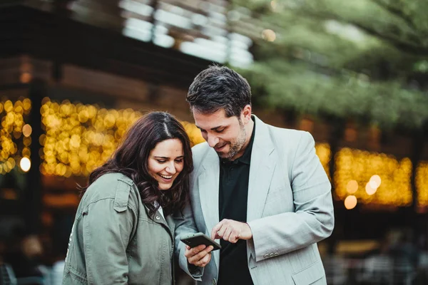 Mutlu bir adam ve kadın, parkta sarı ışıkların olduğu bir cep telefonuna gülümsüyor.