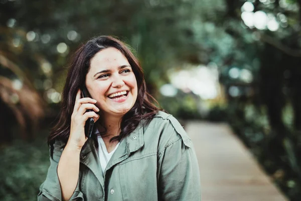 Şehir parkında dikilen genç ve mutlu bir kadın cep telefonuyla gülümsüyor ve sohbeti dinliyor.