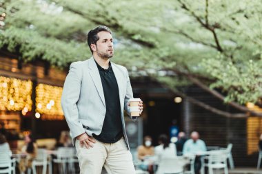 Parkta elinde bir fincan kahveyle sağa bakan dalgın bir iş adamının yatay görüntüsü.