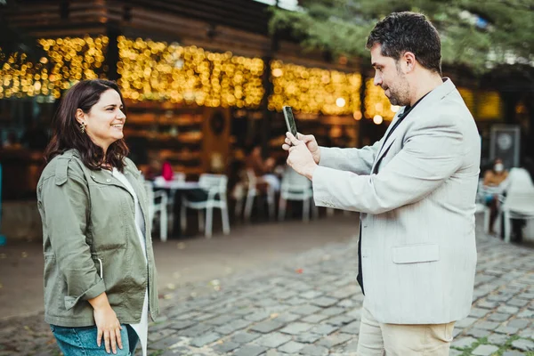 Parkta sarı ışıklı bir kafenin yanındaki bir kadının cep telefonuyla fotoğraf çeken bir adam.