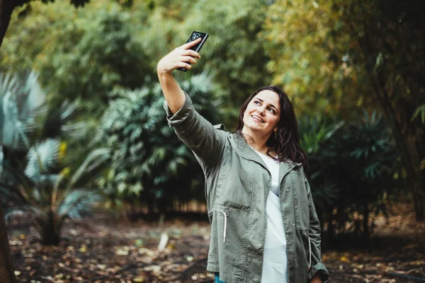 Yeşil çalılarla çevrili bir parkta selfie çeken mutlu bir kadın.