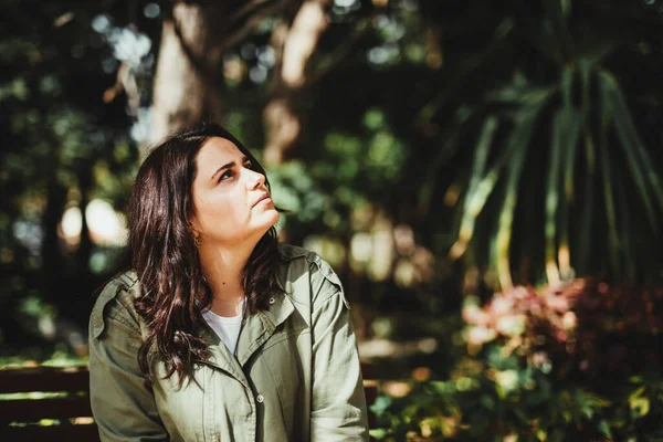 Sağda, şehir parkında kahverengi bir bankta oturan düşünceli bir kadın..