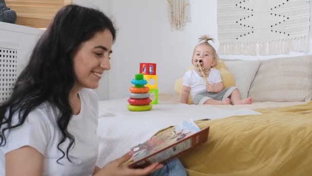 Desarrollo de la primera infancia. Mamá está leyendo un libro para su bebé mientras está sentada en una silla. — Vídeos de Stock