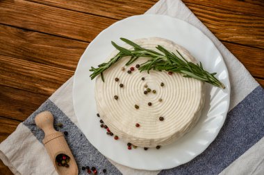 Masada beyaz tabakta bir baş peynir ve yaban mersini ve biberiye var. Taze keçi peyniri ahşap bir masada yatıyor..