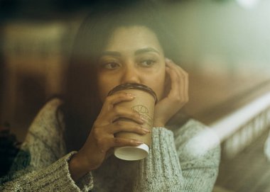 Sokak kafesinin camında yansıma var. Afro-Amerikalı bir kadın elinde bir fincan kahveyle bir kafede oturuyor. Koyu tenli genç bir kadın bir kafede oturur ve ortağını beklerken kahve içer..
