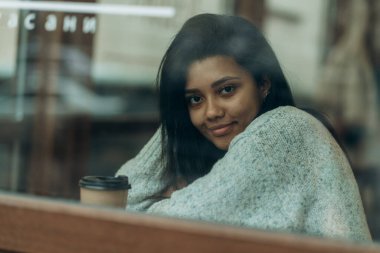 Sokak kafesinin camında yansıma var. Afro-Amerikalı bir kadın elinde bir fincan kahveyle bir kafede oturuyor. Koyu tenli genç bir kadın bir kafede oturur ve ortağını beklerken kahve içer..