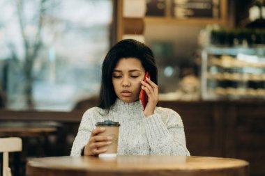 Koyu tenli genç bir kadın bir kafede oturur ve ortağını beklerken kahve içer..