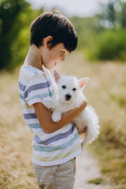 Köpeği olan bir çocuğun arkadaşlığı. Kollarında West Highland White Terrier köpeği tutan mutlu çocuk. Çocuk yazın köpek yavrusuyla parkta yürür. Çocuğun doğadaki en sevdiği köpek..