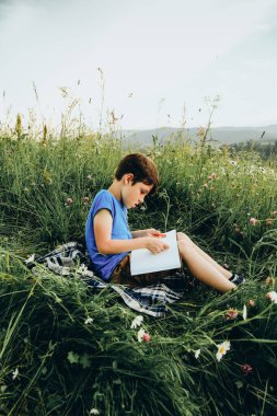 Çocuk dağdaki çimlerin üzerinde oturur ve dağın tepesine bir resim çizer. Genç bir sanatçı yazın bir dağ manzarası çizer..