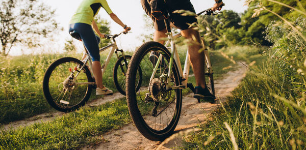 Мужчина и женщина едут на велосипедах по грунтовой дороге летом на закате. Активный отдых летом на велосипедах. Прогулка на семейном велосипеде между полями летом на закате.