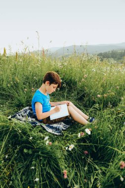 Çocuk dağdaki çimlerin üzerinde oturur ve dağın tepesine bir resim çizer. Genç bir sanatçı yazın bir dağ manzarası çizer..