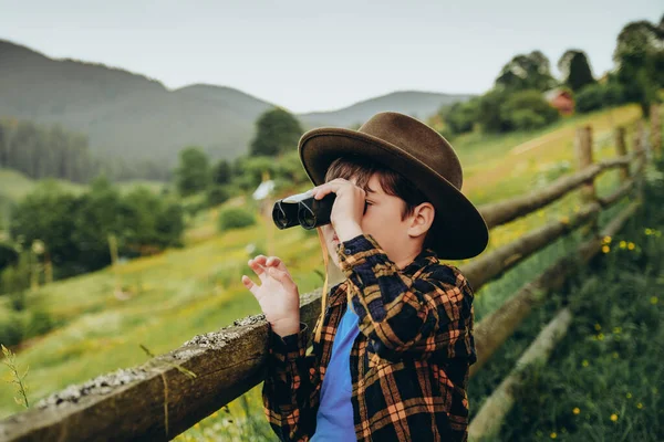 Bir çocuk yazın bir tepede dururken dürbünle bakar. Yazın dağlara aile gezisi. Çocuk yazın dürbünle dağ manzarasını inceliyor..