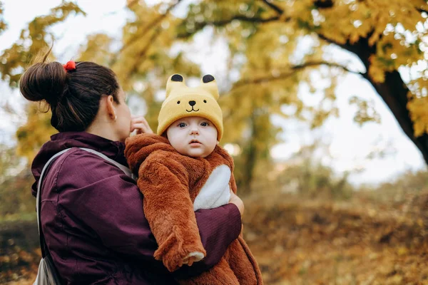 A woman walks in the park in autumn with her child. A young woman holds her baby in her arms and stands under a tree. Family walks in autumn in the park.