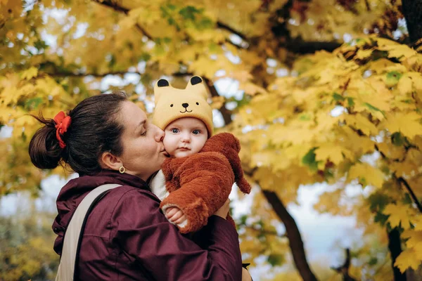 A woman walks in the park in autumn with her child. A young woman holds her baby in her arms and stands under a tree. Family walks in autumn in the park.