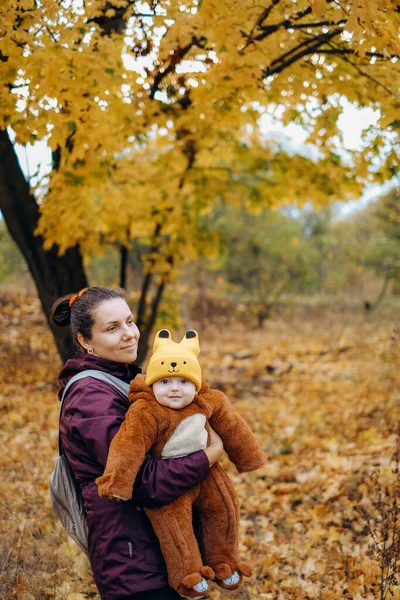 A woman walks in the park in autumn with her child. A young woman holds her baby in her arms and stands under a tree. Family walks in autumn in the park.
