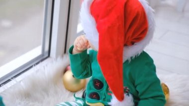 A kid dressed as Santa sits near the window and plays with Christmas decorations in anticipation of the holiday. Evening on the equestrian Christmas.