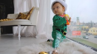 A kid dressed as Santa sits near the window and plays with Christmas decorations in anticipation of the holiday. Evening on the equestrian Christmas.