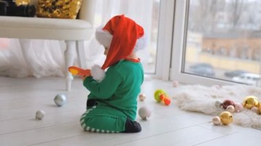 A kid dressed as Santa sits near the window and plays with Christmas decorations in anticipation of the holiday. Evening on the equestrian Christmas.