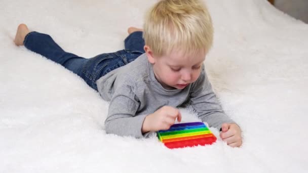 El niño miente y juega un juego educativo. Juguete sensorial colorido antiestrés nervioso empuje pop en las manos de los niños pequeños. Antiestrés de moda pop it toy. Arco iris nerviosa sensorial. Juguete de silicona de moda — Vídeos de Stock