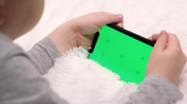 Anaokulunda elinde aletle cep telefonundan video izleyen bir çocuk. Çocuk elinde akıllı telefon yeşil ekranı kullanıyor ve tutuyor. Teknoloji üretimi konsepti. Cep telefonunun krom anahtarına odaklan