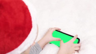 Noel Baba şapkalı çocuk evde cep telefonuyla video oyunu oynuyor. Çocuk elinde akıllı telefon yeşil ekranı kullanıyor ve tutuyor, Noel konsepti. Kromakey 'e odaklan