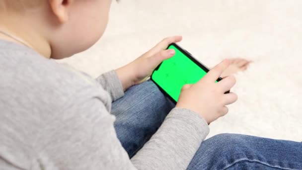 Niño preescolar con gadget jugando videojuegos digitales en el teléfono móvil en casa. Niño usando y sosteniendo una pantalla verde smartphone en la mano, concepto de generación de tecnología. Concéntrate en chromakey — Vídeos de Stock