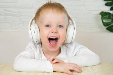 Küçük Avrupalı çocuk büyük beyaz kulaklıklı beyaz bir tişörtle gülümsüyor, müzik dinliyor ve kameraya bakıyor. Ağır çekim. Müzik konsepti, ev eğlencesi. 4k. 