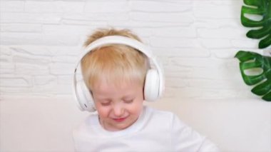 Küçük Avrupalı bir çocuk beyaz bir tişörtle büyük beyaz kulaklıklarla gülümsüyor ve gözleri kapalı aktif olarak dans ediyor ve müzik dinliyor. Ağır çekim. Müzik konsepti, ev eğlencesi. 4k 