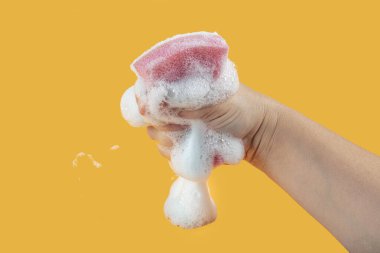 Elde bulaşık yıkamak için sünger. Temizlik süngeri tutuyor ve sarı arka planda sabunlu köpüğü siliyor. Temizlik konsepti, temizlik hizmeti. Pankart. Düz yatış, üst görünüm.