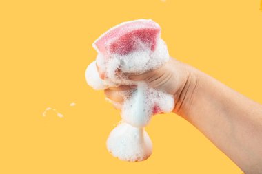 Elde bulaşık yıkamak için sünger. Temizlik süngeri tutuyor ve sarı arka planda sabunlu köpüğü siliyor. Temizlik konsepti, temizlik hizmeti. Pankart. Düz yatış, üst görünüm.