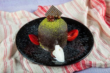 Single sweet cake. Glazed chocolate cake, one piece, on black plate with pistachio cake. It is served on a plate with ice cream and strawberries.