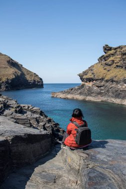 Kadın kayaların üzerinde oturuyor, arkası dönük ve önüne bakıyor. Cornwall, Boscastle, İngiltere 'de güzel okyanus manzarası.
