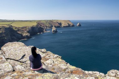 Kadın kayaların üzerinde oturuyor, arkası dönük ve önüne bakıyor. Cornwall, Boscastle, İngiltere 'de güzel okyanus manzarası.