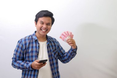 Mavi ekose gömlekli heyecanlı Asyalı adamın portresi beyaz arka planda duruyor, cep telefonu kullanırken yüz bin rupiayı gösteriyor. Mali ve tasarruf kavramı.