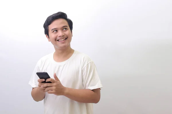 一个身穿白色T恤的兴奋的亚洲男人的画像 他看着什么 拿着手机 白色背景上的孤立图像 广告概念 — 图库照片