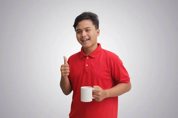 穿着红色马球衫 站在灰蒙蒙的背景下 拿着一杯咖啡 用大拇指举起手势微笑的亚裔男子的画像 — 图库照片