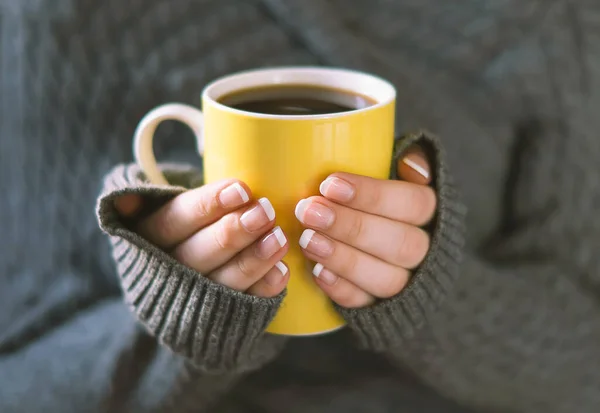 A girl in a warm knitted cardigan holds a yellow mug with a hot drink in her hands and warms up. Beautiful hands of a young girl holding a cup of black coffee, close-up. The concept of comfort, warmth