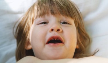 Güzel, güzel, şaşırmış küçük sarışın kız kanepede uzanıp kameraya bakıyor. Yapraklı dişleri söktükten sonra bebek. Gözlere odaklan.