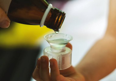 Bir çocuk eli öksürük şurubu için yakın plan bir ölçü kabı tutar. Anne şişeden kaşığa ilaç ya da antipiretik döker. Sağlık, insan ve ilaç kavramı.