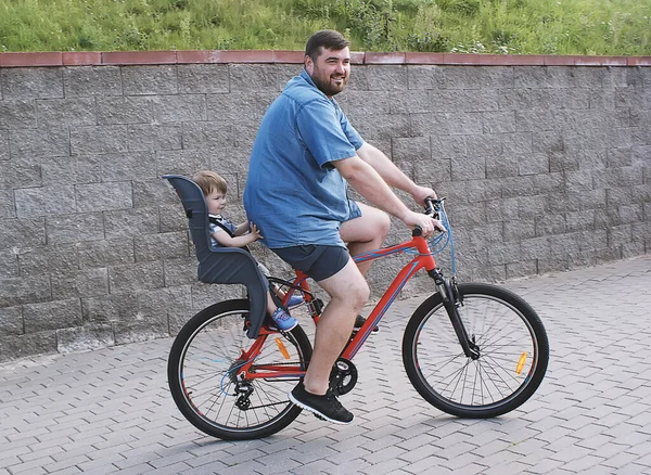 Bir baba, küçük kızı güneşli bir günde güvenli bir çocuk koltuğunda otururken bisiklete biner. Bisikletli bir adam çocukla bisiklet sürüyor, bisiklet koltuğunda, yan manzaralı. Aile tatili kavramı.