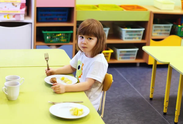 かわいい赤ちゃんの女の子は幼稚園で健康的な食べ物を食べている かなり小さな女の子がテーブルに座って昼食を取っている かわいい子供は子供の教育センターでカツとジャガイモを食べる ロイヤリティフリーのストック画像