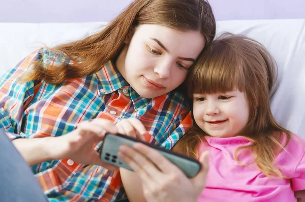 幸せな姉妹のソファに一緒に携帯電話で漫画を見て座っている 姉はかわいい妹にスマートフォンの画面上で面白いビデオをオンラインで示しています ガジェットと子供たち ストック写真