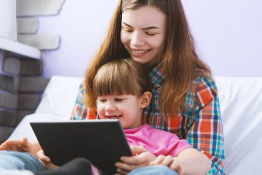 İki tatlı kız kardeş kanepede oturup tablet bilgisayarda çizgi film izliyorlar. Abla tablet ekranında komik kız kardeş eğitim oyunlarını gösteriyor..