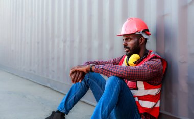 Afrikalı Amerikalı bir erkek teknisyen ya da mühendis portresi. Konteynırın yanına otur ve yorgun, uykulu ya da işsiz görün. Lojistik endüstriyel kargo taşıyıcısı konsepti. İthalat-ihracat ticari ulaşım